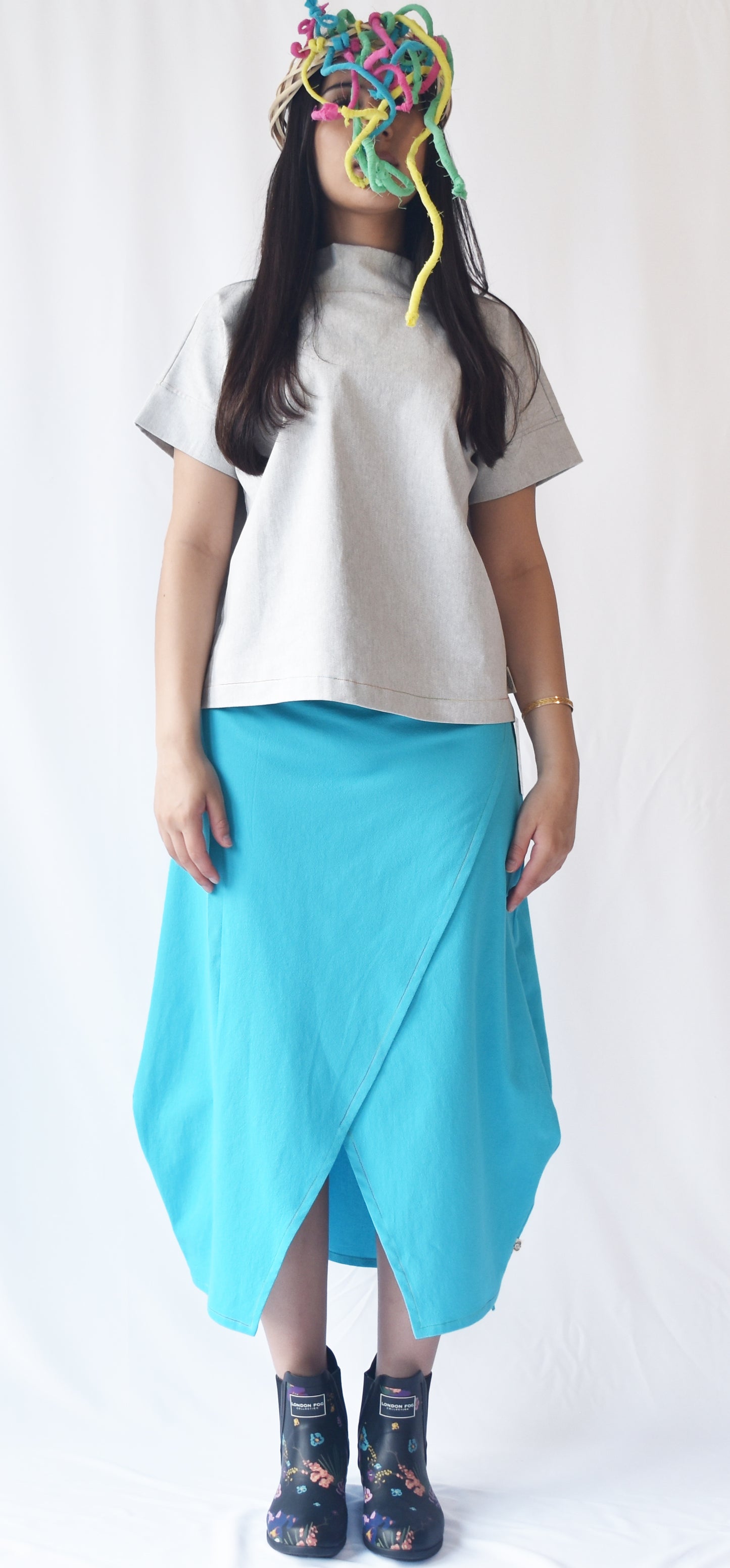 Woman wearing a blue wrap-style midi skirt with grey top and printed ankle boots. The SheKnows Brand skirt features an adjustable drape and midi length.
