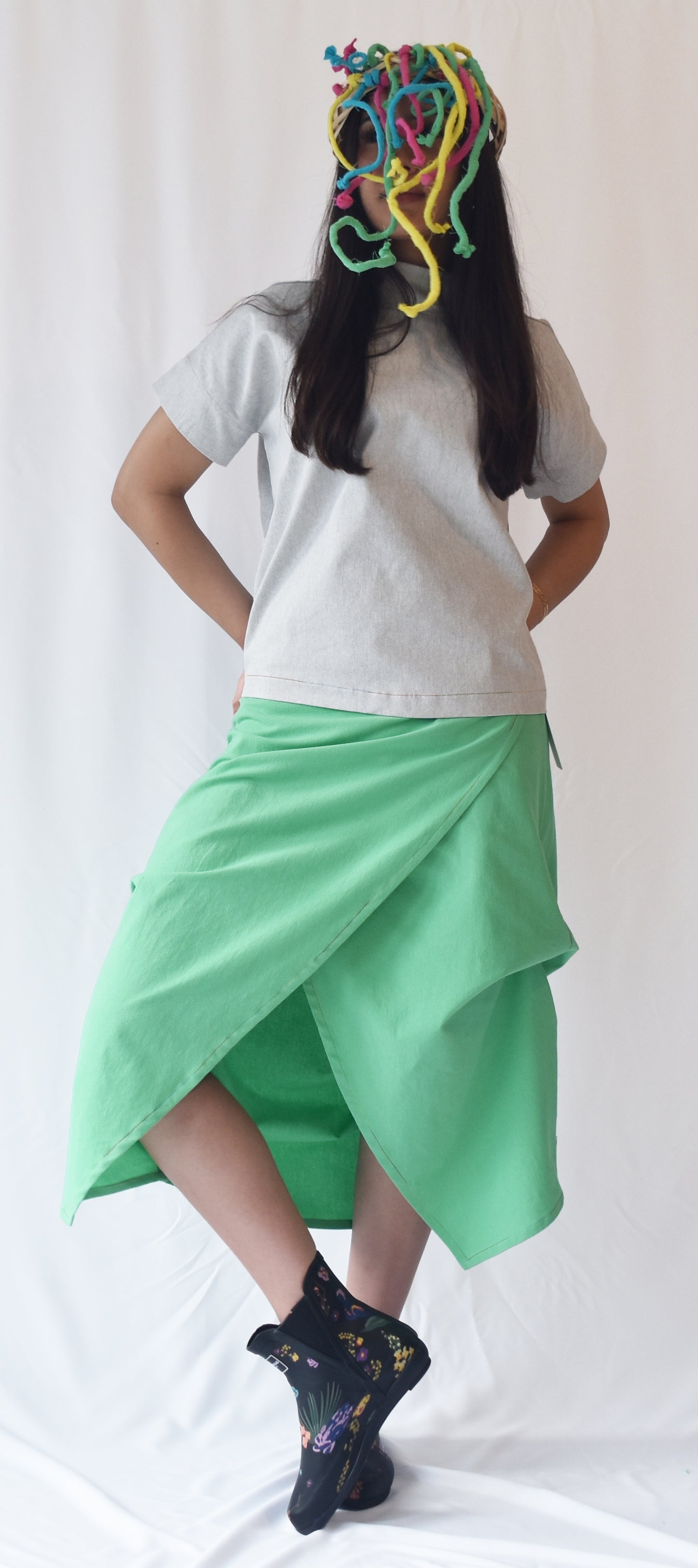 Woman wearing a green wrap-style midi skirt with grey top and printed ankle boots. The SheKnows Brand skirt features an adjustable drape and midi length.