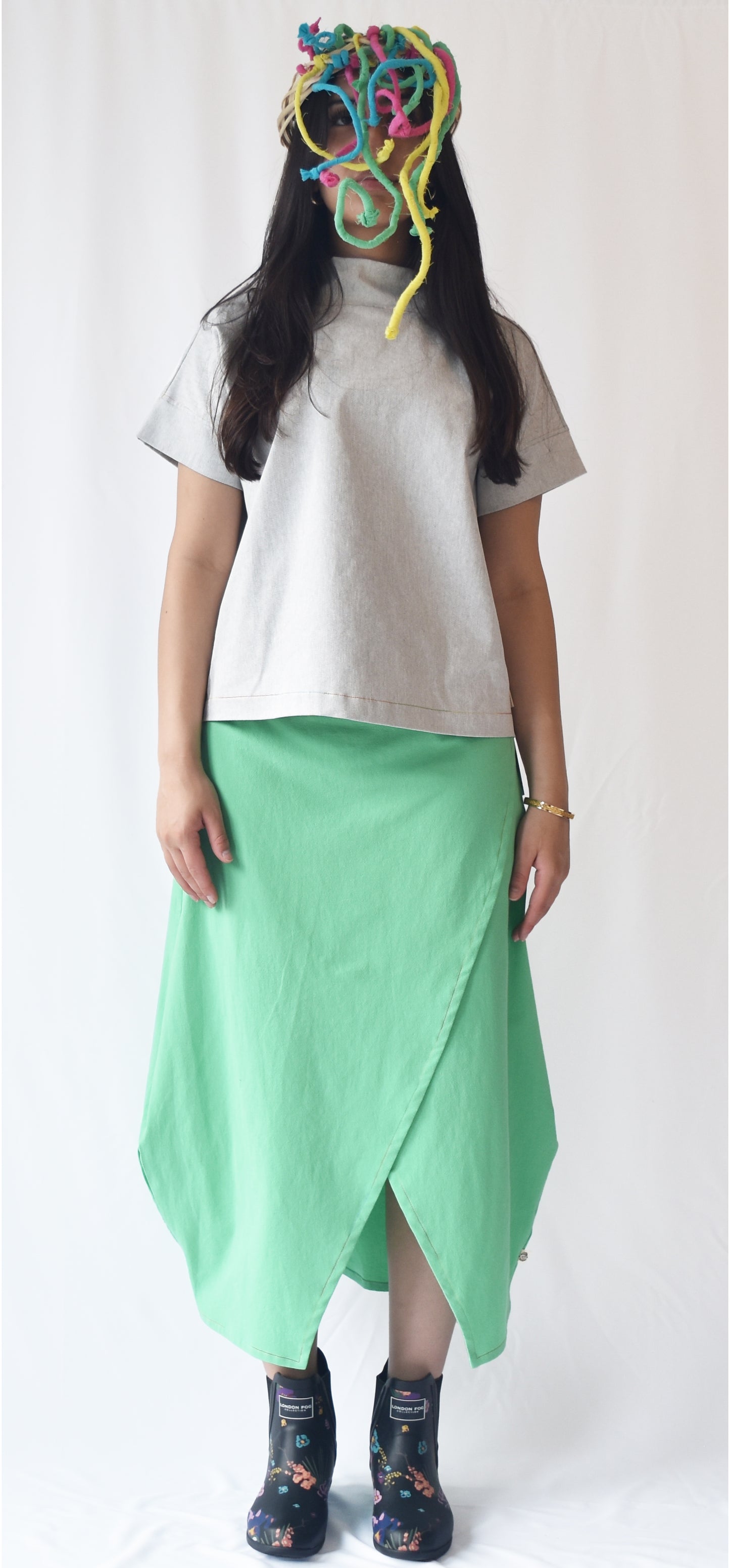 Woman wearing a green wrap-style midi skirt with grey top and printed ankle boots. The SheKnows Brand skirt features an adjustable drape and midi length.