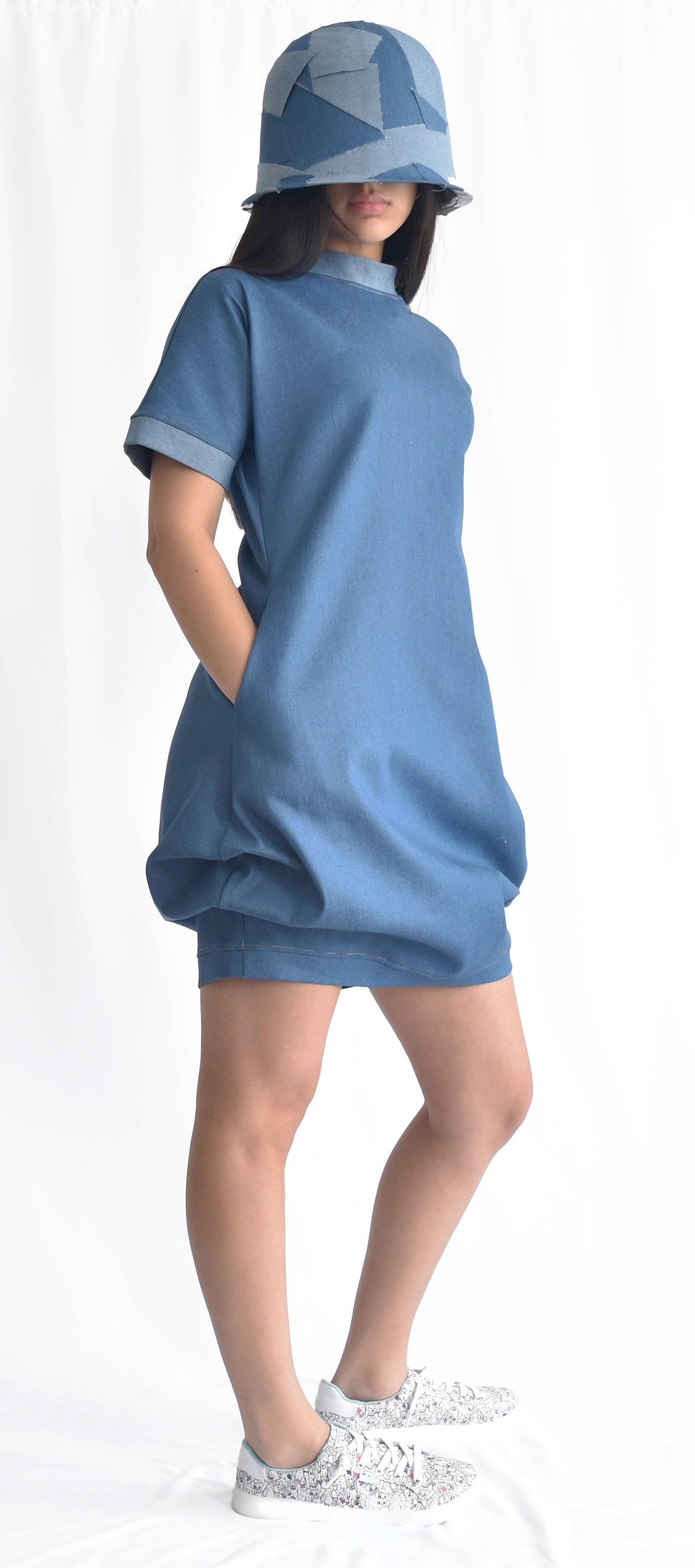 Woman wearing a short medium blue denim dress with contrast light blue stand collar and cuffs on short sleeves. The SheKnows Brand dress has pockets and a structured and playful silhouette