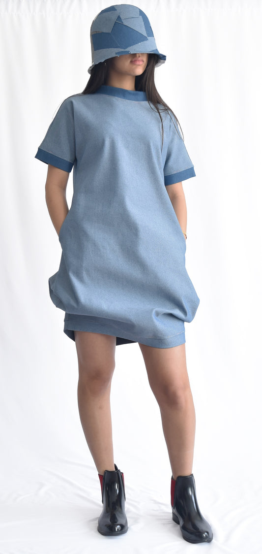 Woman wearing a short light blue denim dress with contrast medium blue stand collar and cuffs on short sleeves. The SheKnows Brand dress has pockets and a structured and playful silhouette