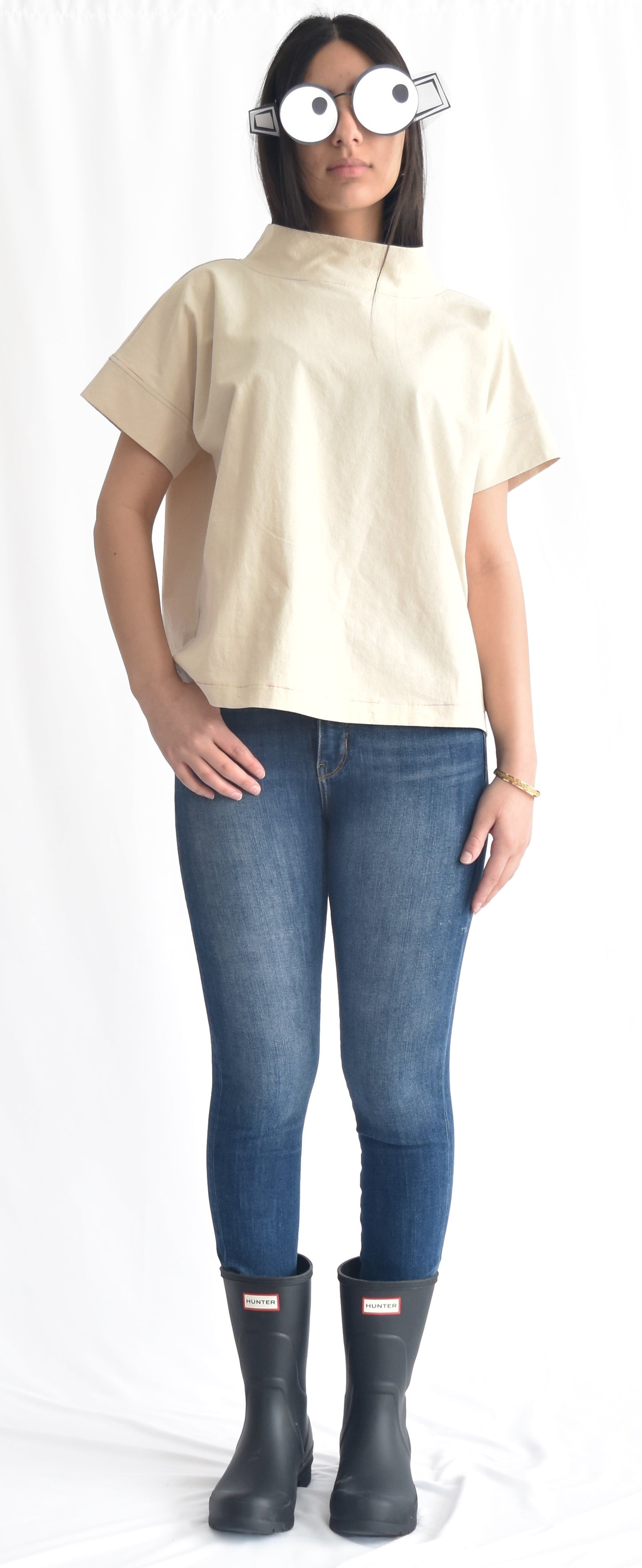 Woman wearing a minimalist cotton top with short sleeves and stand collar in birch from SheKnows Brand. The top has a high-hip length and relaxed fit.
