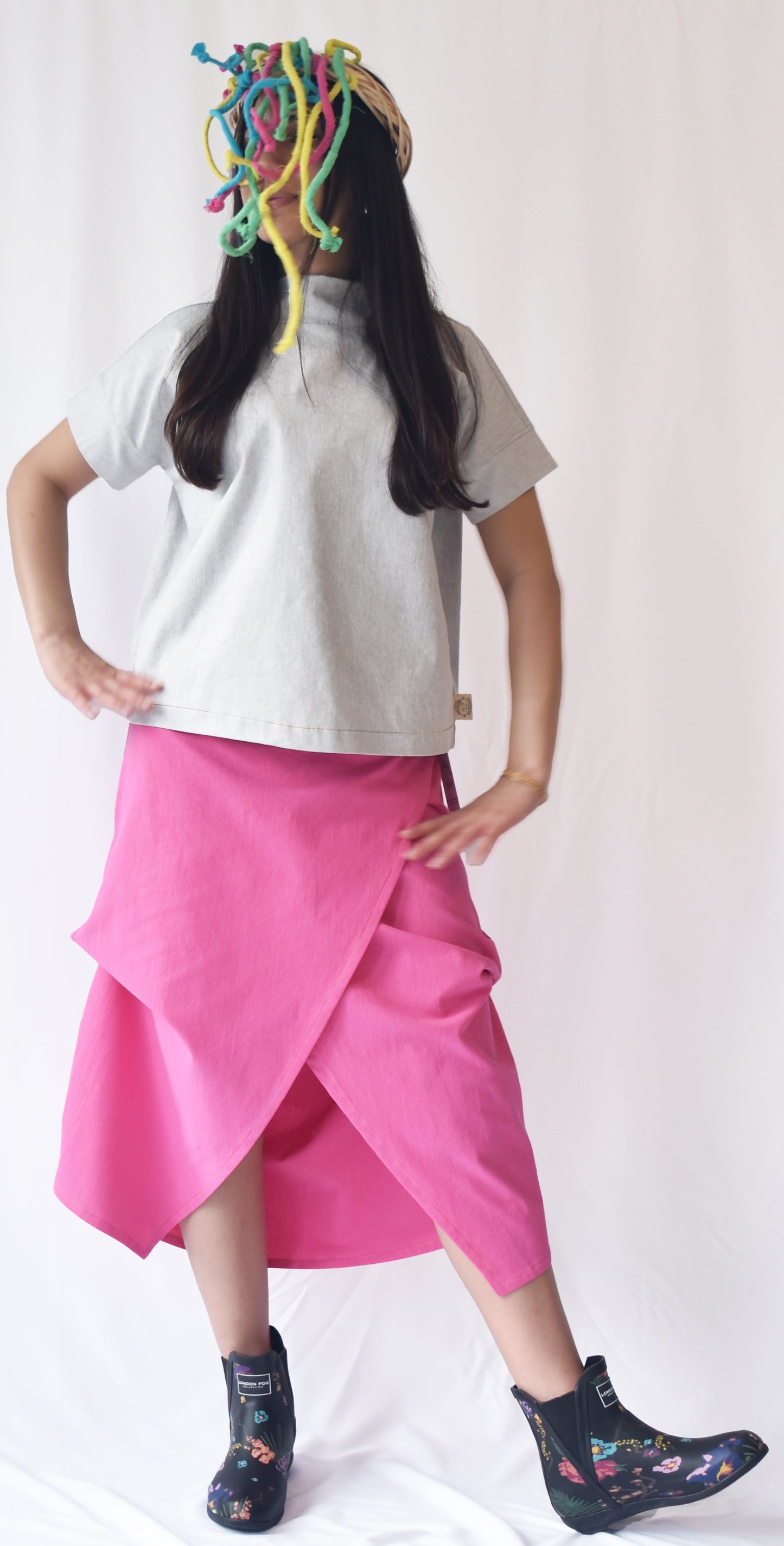 Woman wearing a pink wrap-style midi skirt with grey top and printed ankle boots. The SheKnows Brand skirt features an adjustable drape and midi length.