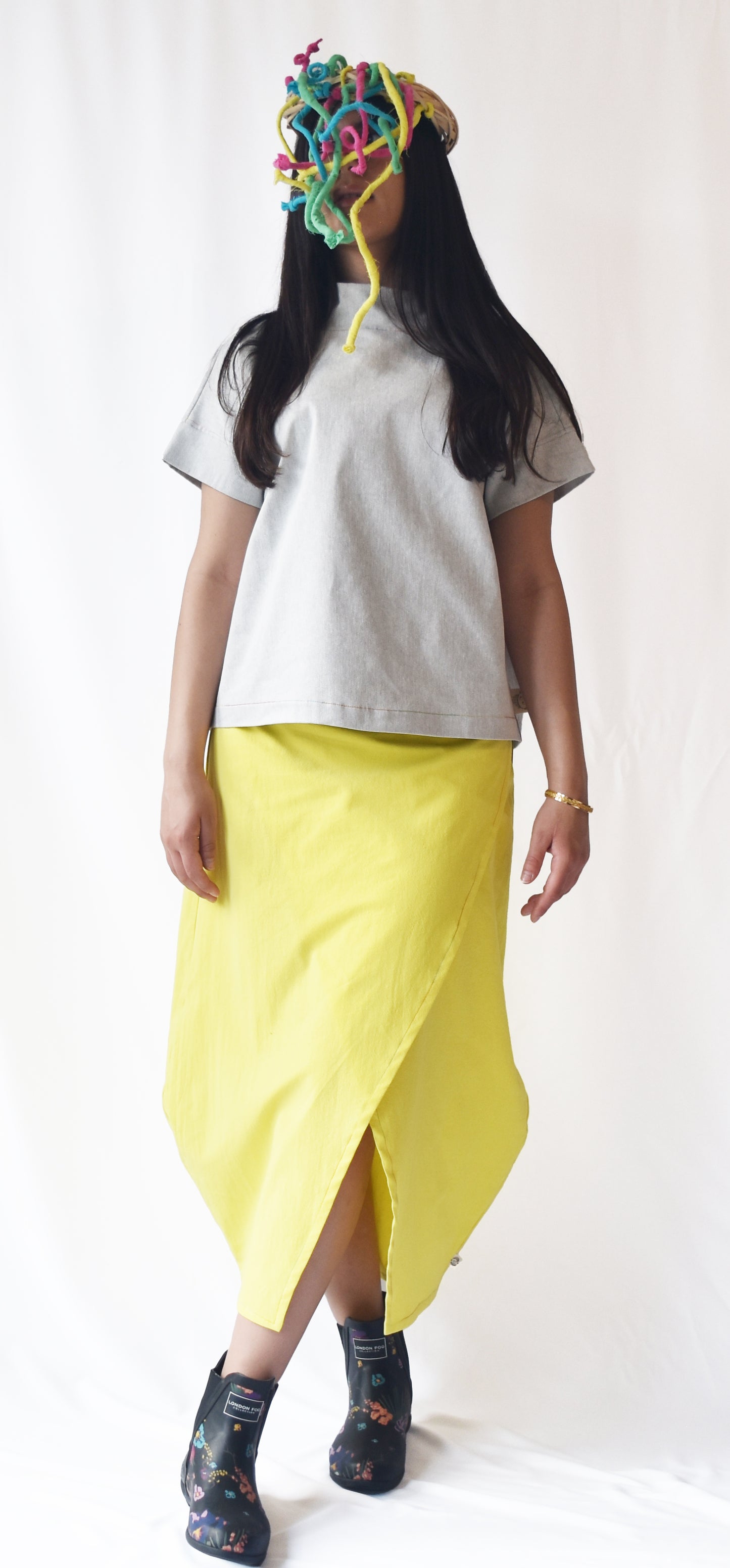 Woman wearing a yellow wrap-style midi skirt with grey top and printed ankle boots. The SheKnows Brand skirt features an adjustable drape and midi length.