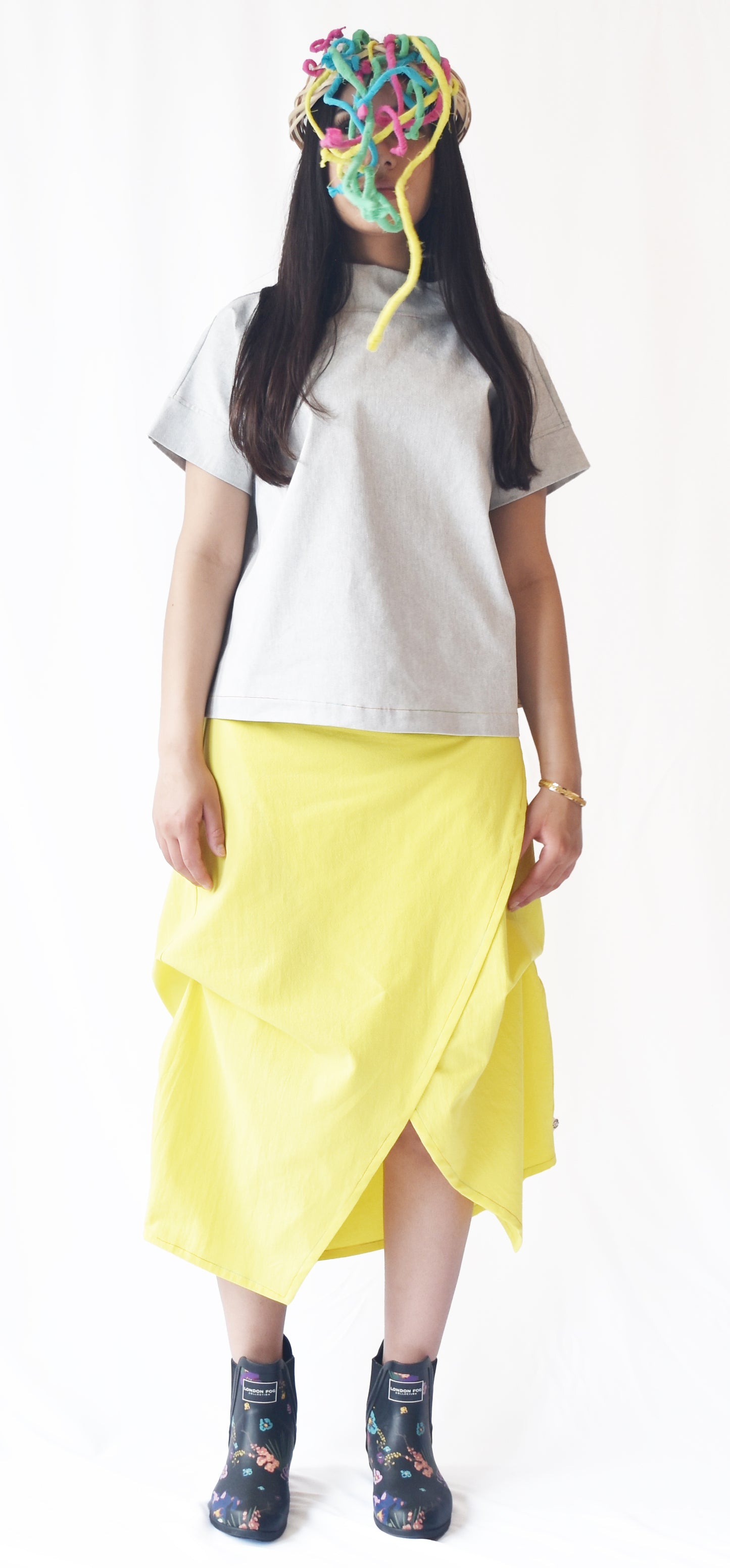 Woman wearing a yellow wrap-style midi skirt with grey top and printed ankle boots. The SheKnows Brand skirt features an adjustable drape and midi length.