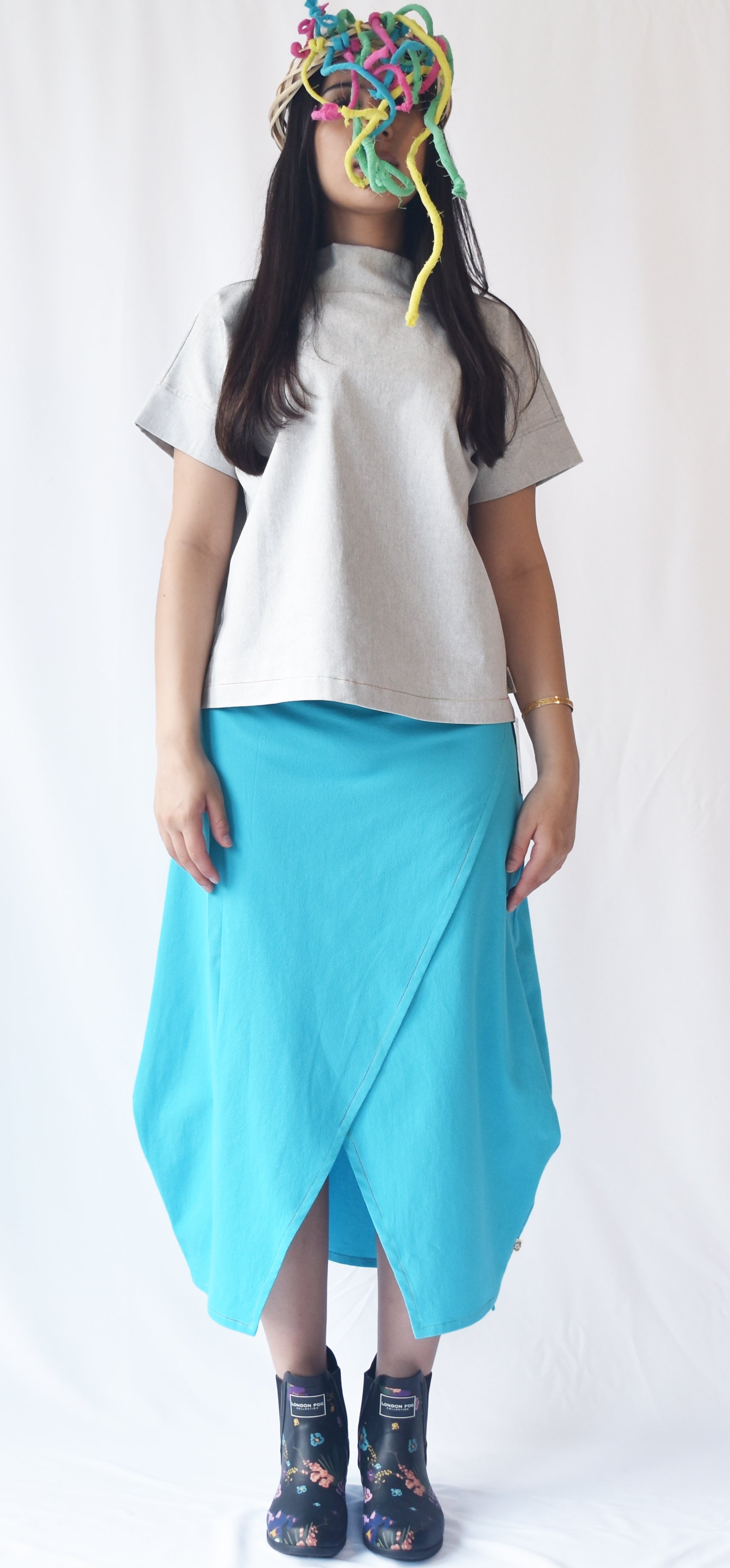 Woman wearing a blue wrap-style midi skirt with grey top and printed ankle boots. The SheKnows Brand skirt features an adjustable drape and midi length.