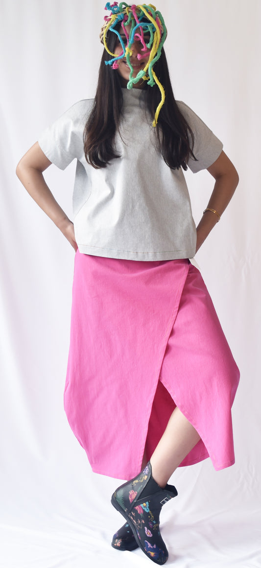 Woman wearing a pink wrap-style midi skirt with grey top and printed ankle boots. The SheKnows Brand skirt features an adjustable drape and midi length.
